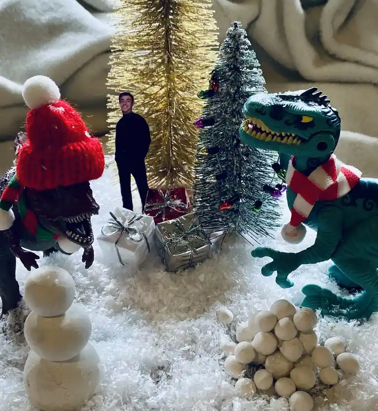 Two toy dinosaurs dressed in Christmas sweaters and scarves stand in a snowy scene with miniature Christmas trees, wrapped…