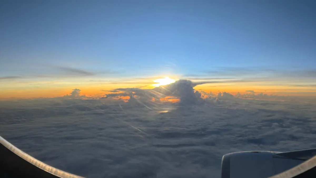 A view from an airplane window shows a sunrise or sunset over a sea of clouds, with the sun partially obscured by a large…