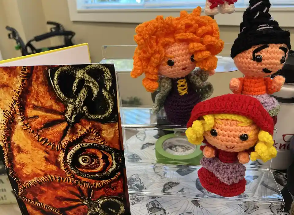 Three crocheted dolls resembling characters from 'Hocus Pocus' sit on a clear acrylic shelf next to a book with a dark…