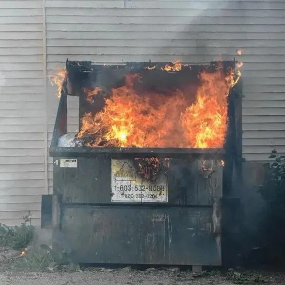 A large, dark green dumpster is engulfed in bright orange flames and billowing smoke, positioned against a light-colored…