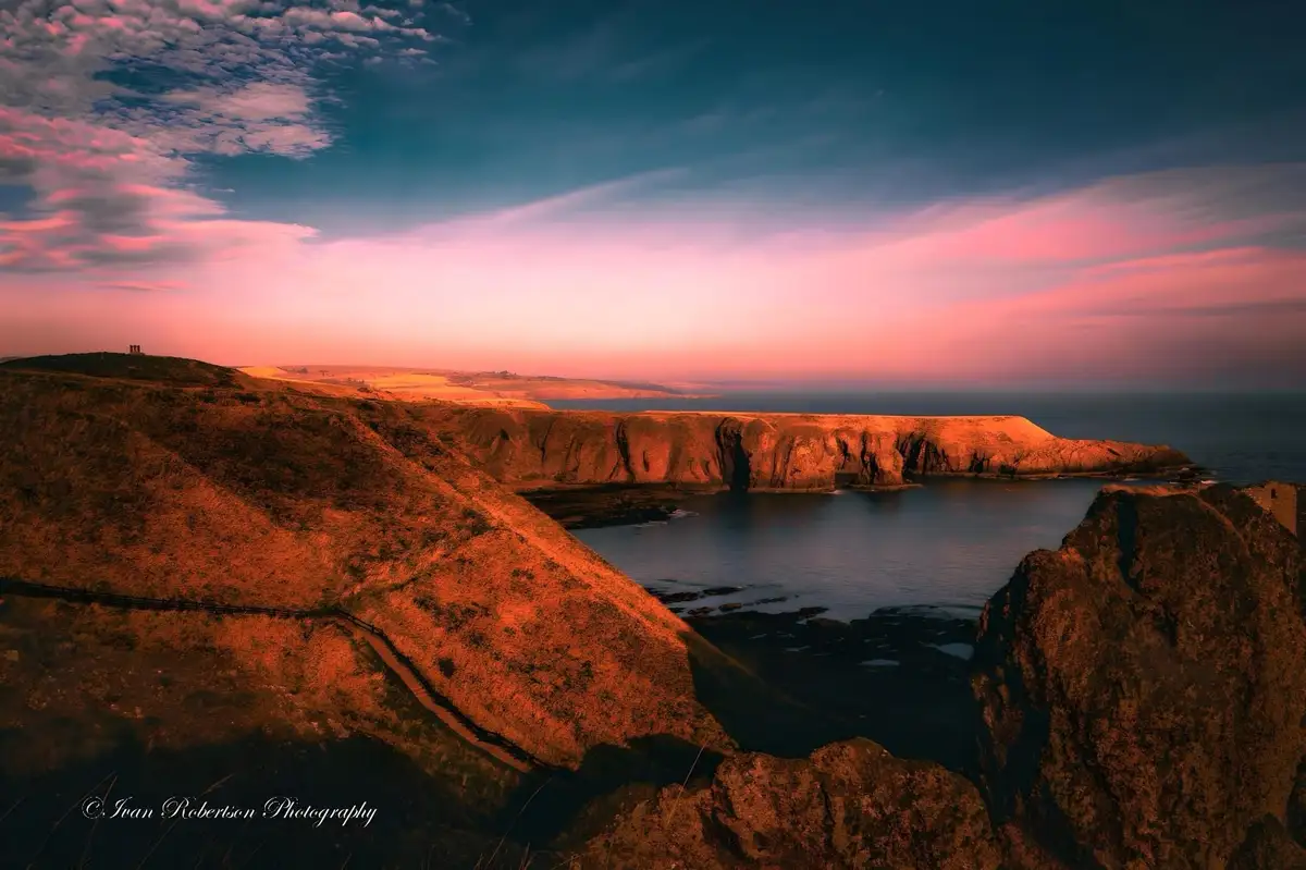 A scenic photograph of a rugged coastal cliffside at sunset, with vibrant pink and orange hues in the sky and on the cliffs…