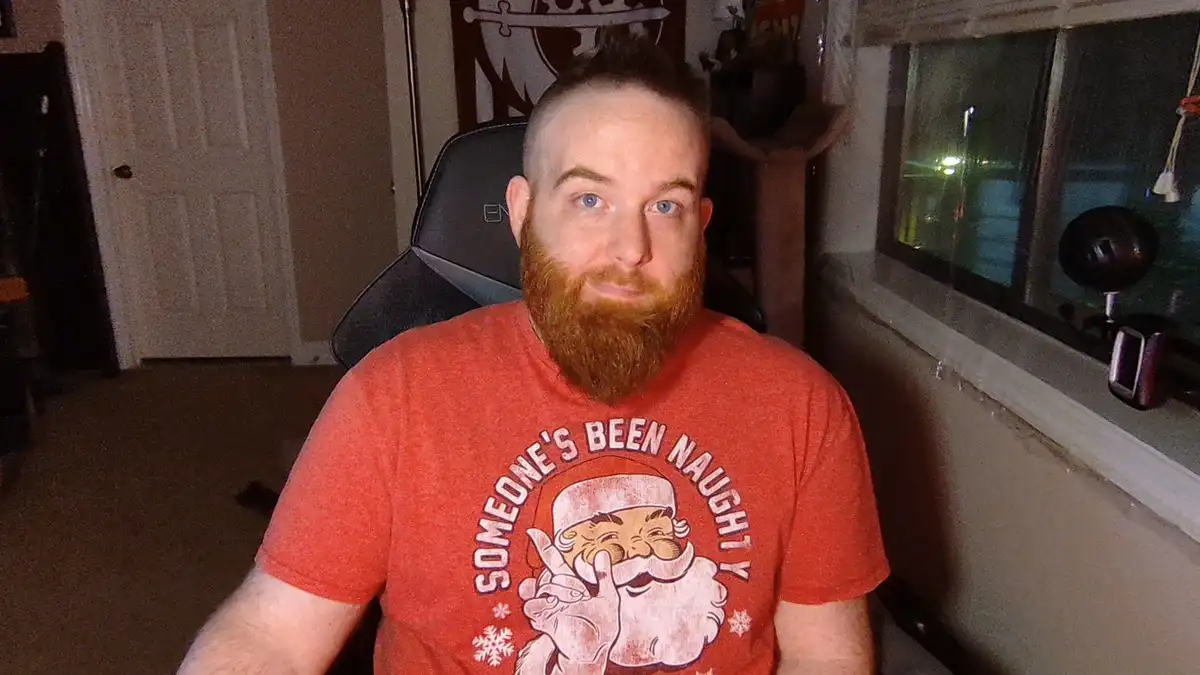 A man with a red beard and blue eyes sits in a gaming chair wearing a red t-shirt that says 'SOMEONE'S BEEN NAUGHTY!' with a…