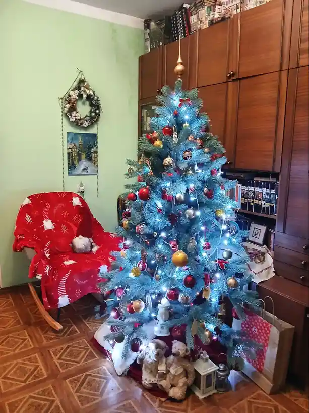 a brightly lit, blue-tinted artificial christmas tree decorated with red and gold ornaments stands in a room with wooden…