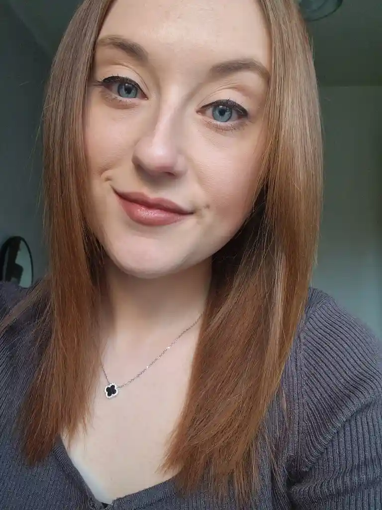 A close-up selfie of a woman with long brown hair and blue eyes, wearing a grey ribbed sweater and a silver necklace with a…