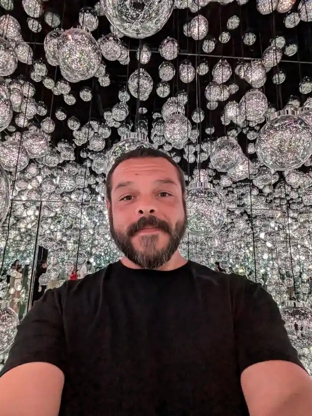 A man with a beard and black t-shirt takes a selfie in front of a dazzling installation of thousands of hanging, glittery…