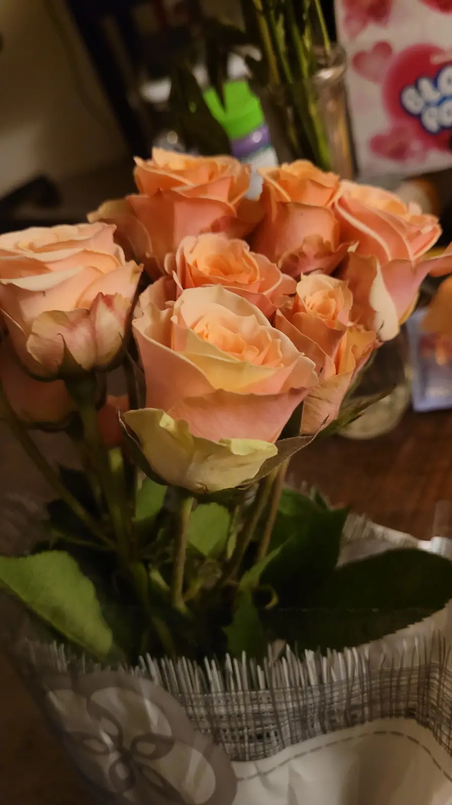 A close-up photo shows a bouquet of peach-colored roses in a clear glass vase, some petals slightly wilted. The background…