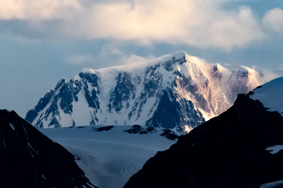 A majestic, snow-covered mountain range is bathed in soft, golden light breaking through clouds, framed by dark silhouetted…