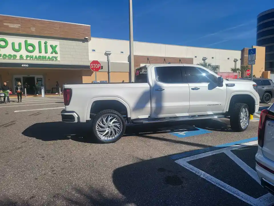 A white Chevrolet Silverado Denali pickup truck with large custom wheels is parked in a blue handicapped parking space in…
