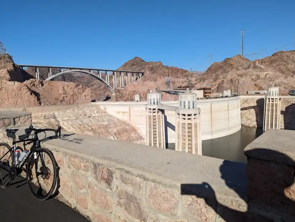 A black road bicycle leans against a stone railing overlooking the Hoover Dam and the Colorado River under a clear blue sky…