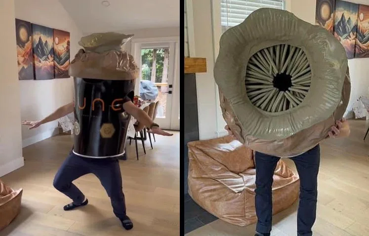 A person in a homemade costume of a black 'JUNE' branded coffee cup with a plastic lid stands in a living room, arms…