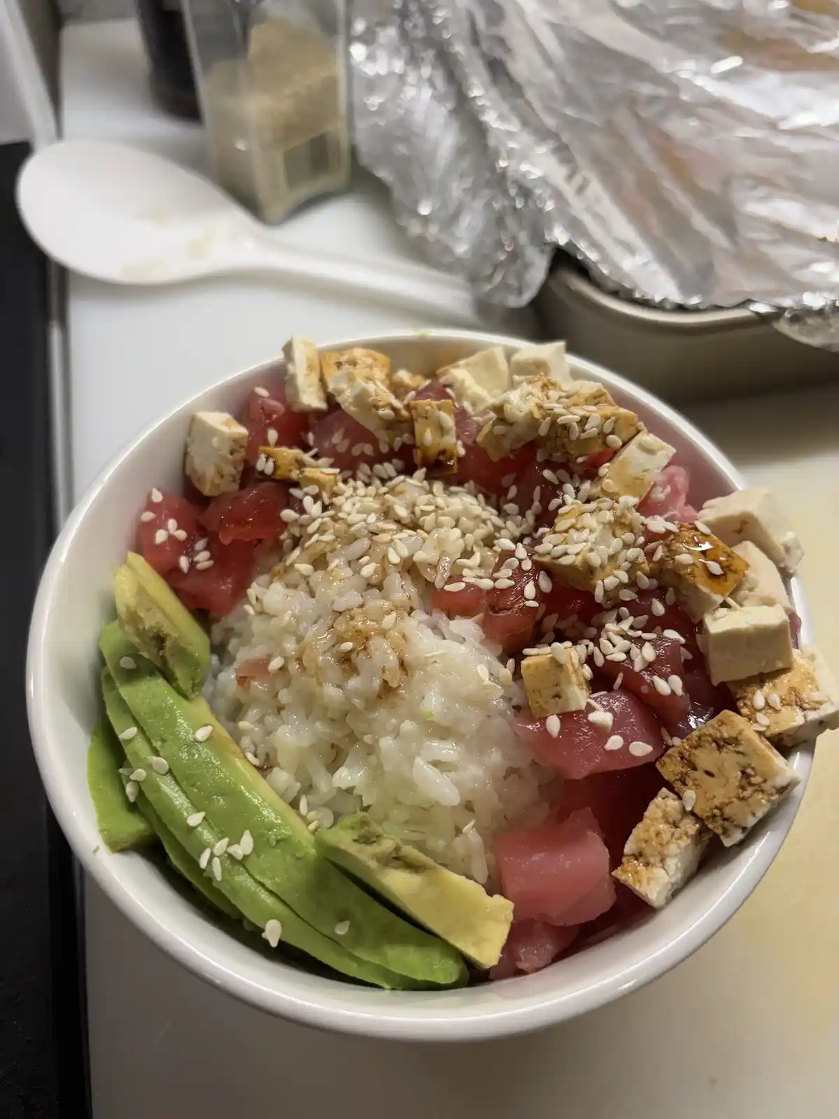 A close-up photo of a white bowl filled with rice, diced raw tuna, cubed tofu, sliced avocado, and white sesame seeds. In…