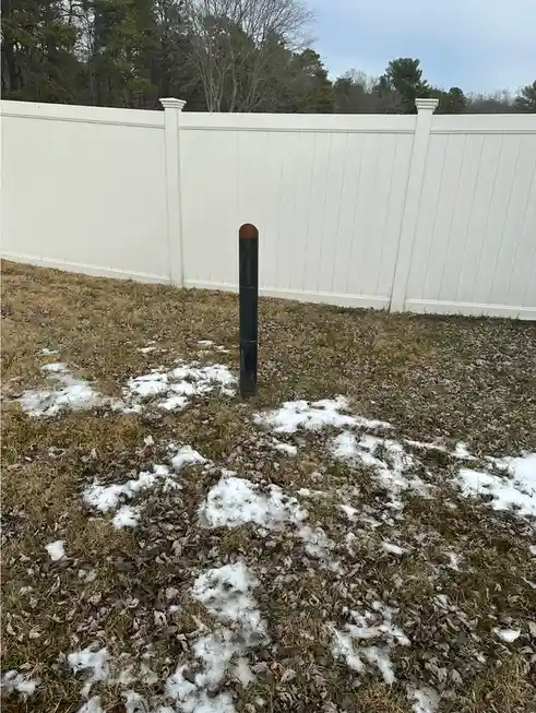A black cylindrical pole stands upright in a grassy yard with patches of melting snow, in front of a white vinyl fence. The…