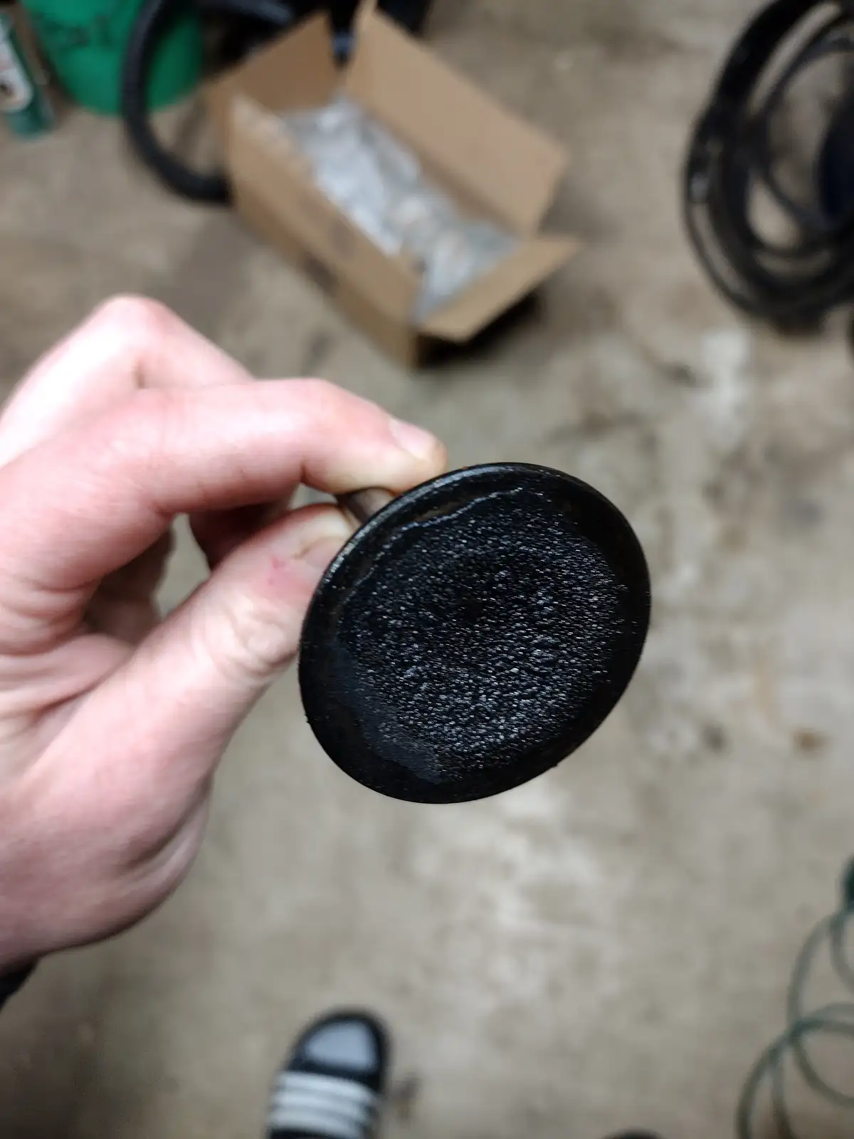 A hand holds a dirty, black, circular carburetor part covered in grime and residue, with a blurry garage background showing…
