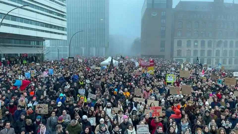 A large crowd of protesters fills a city street, holding various handmade signs and banners. Many people are bundled up in…