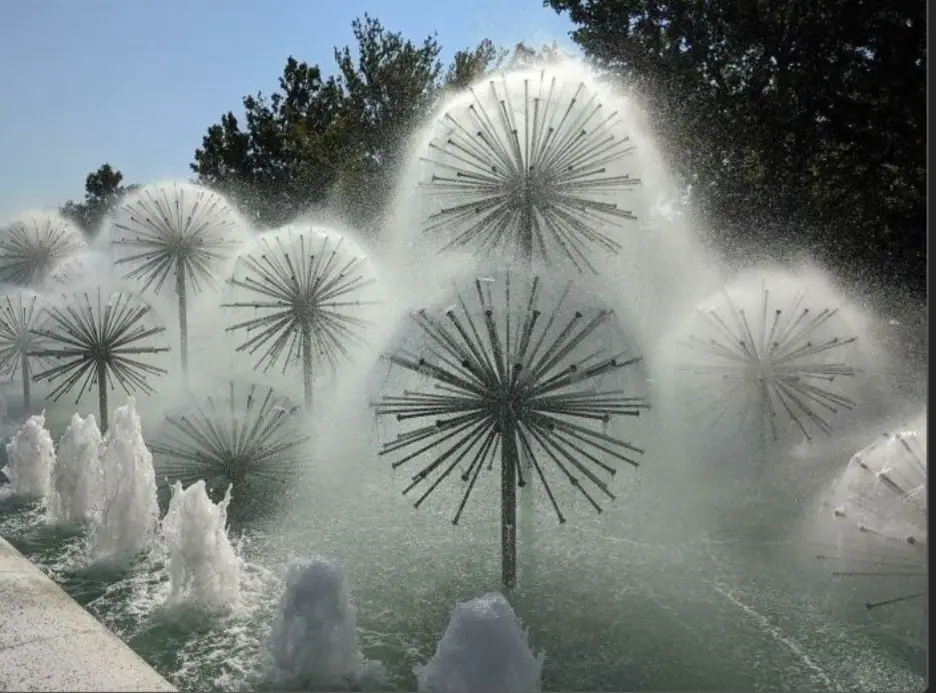 A photo of a fountain with multiple large, starburst-shaped water jets resembling dandelion clocks, set against a backdrop…