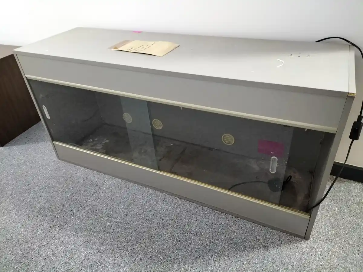 A grey, empty, glass-fronted cabinet sits on a grey carpeted floor, with a small handwritten note on top and a black power…