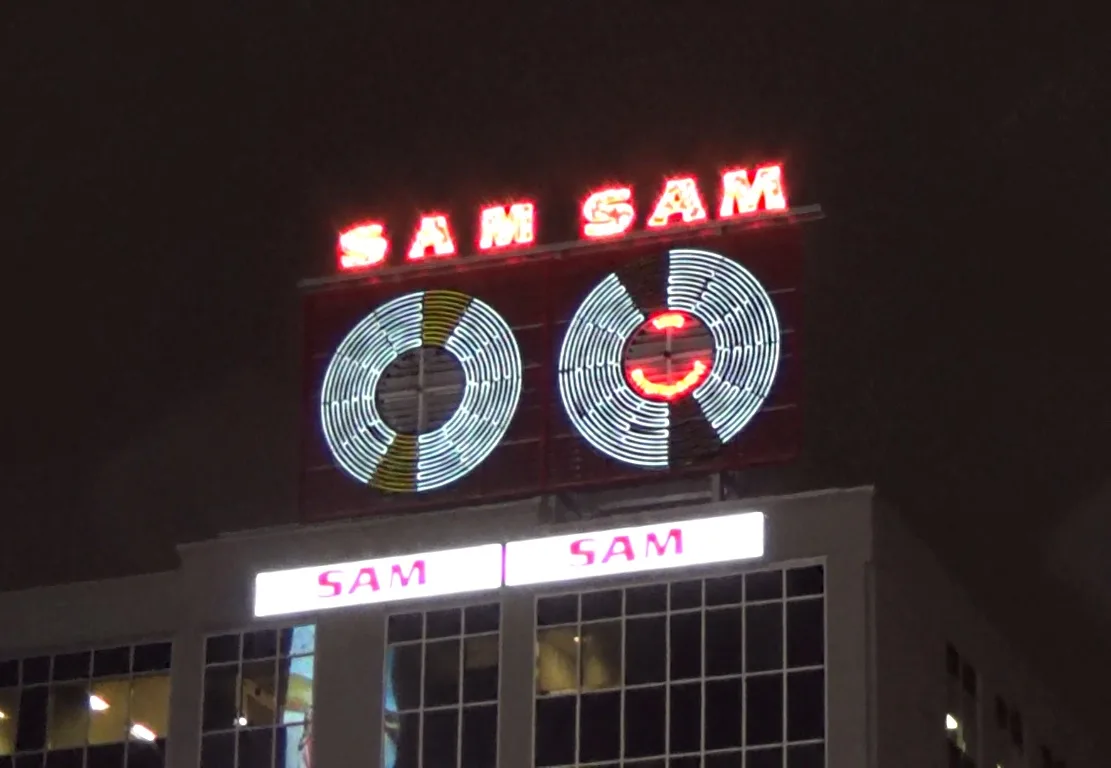 A nighttime photo of a building's illuminated sign that reads 'SAM SAM' in red neon letters above two large circular…
