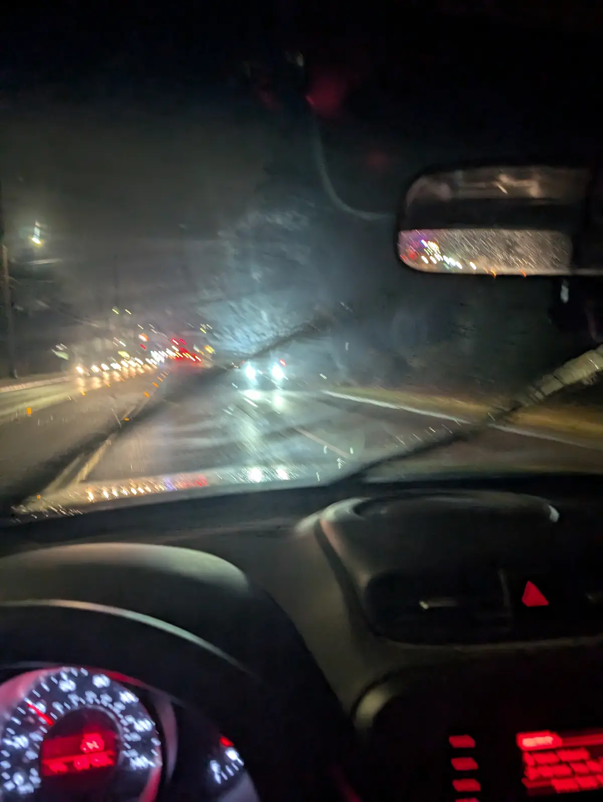 A blurry nighttime photo taken from inside a car, showing a wet windshield with streaks from windshield wipers, illuminated…