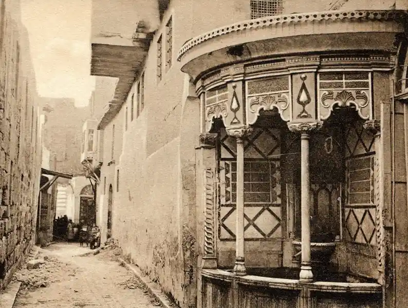 this is a sepia-toned historical photograph of a narrow alleyway in an old city, featuring a prominent ornate wooden balcony…