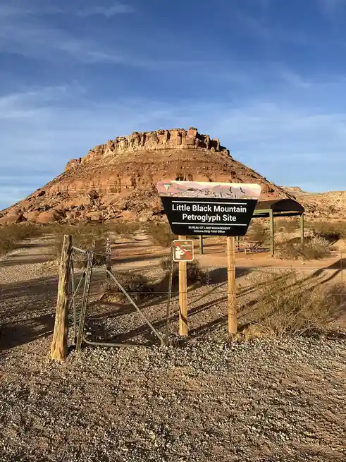 A desert landscape features a large, reddish-brown rock formation behind a wooden sign that reads 'Little Black Mountain…