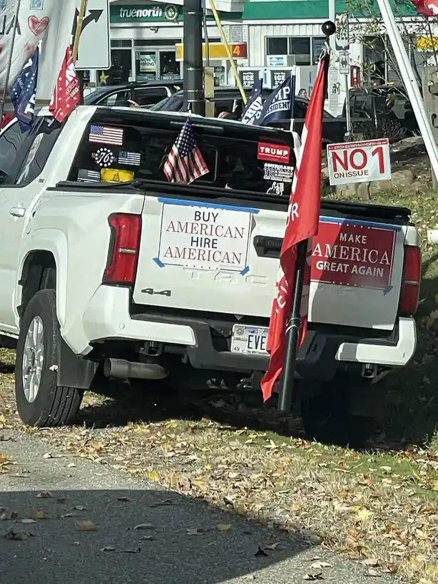 A white pickup truck is parked outdoors with autumn leaves on the ground, covered in political and patriotic signs. Visible…