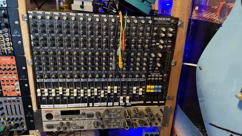 A close-up photo of a densely packed audio mixing console with numerous knobs, faders, and patch cables, surrounded by other…