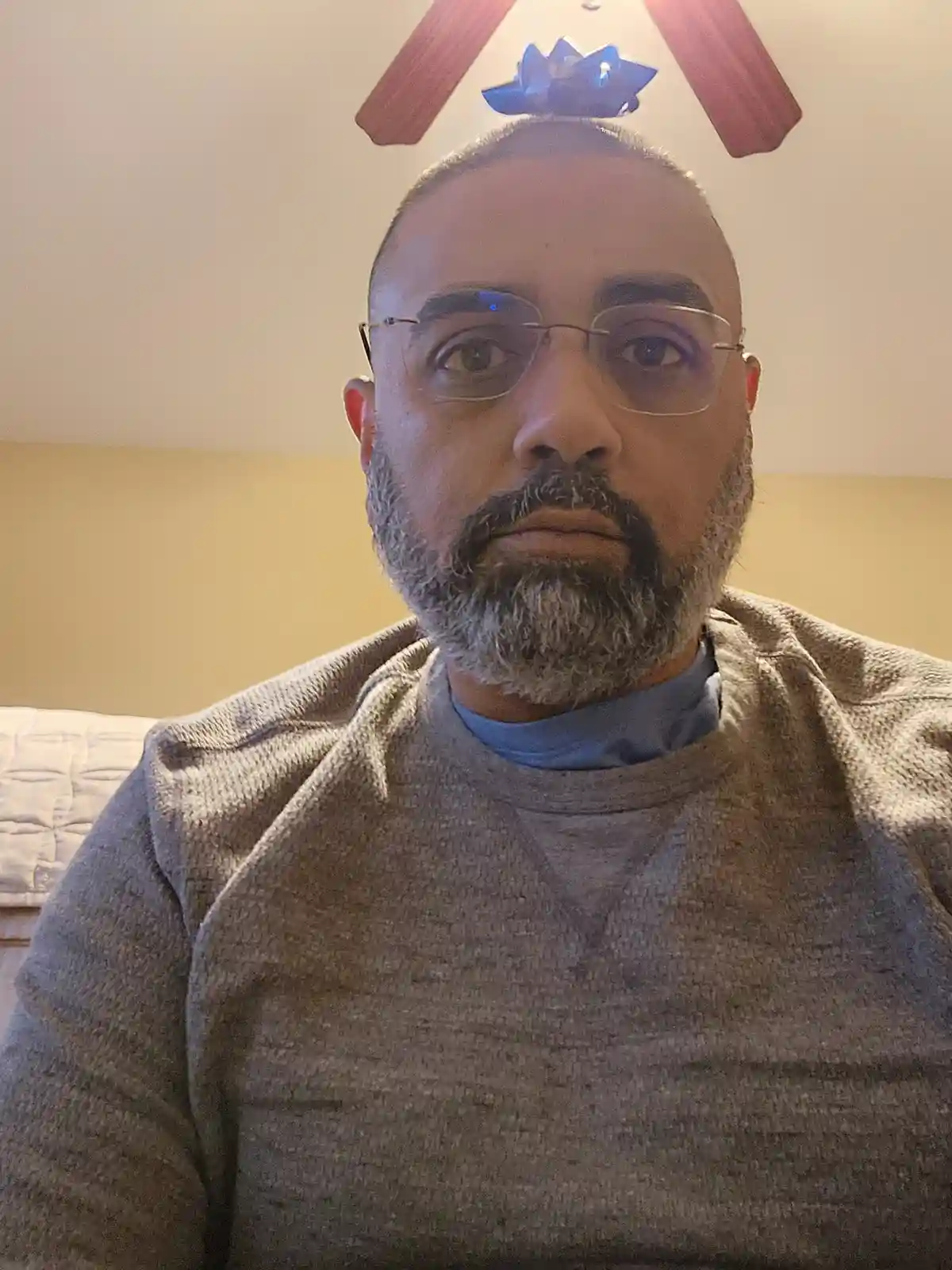 A man with glasses and a gray beard stares directly at the camera with a neutral expression. Above his head, a ceiling fan…