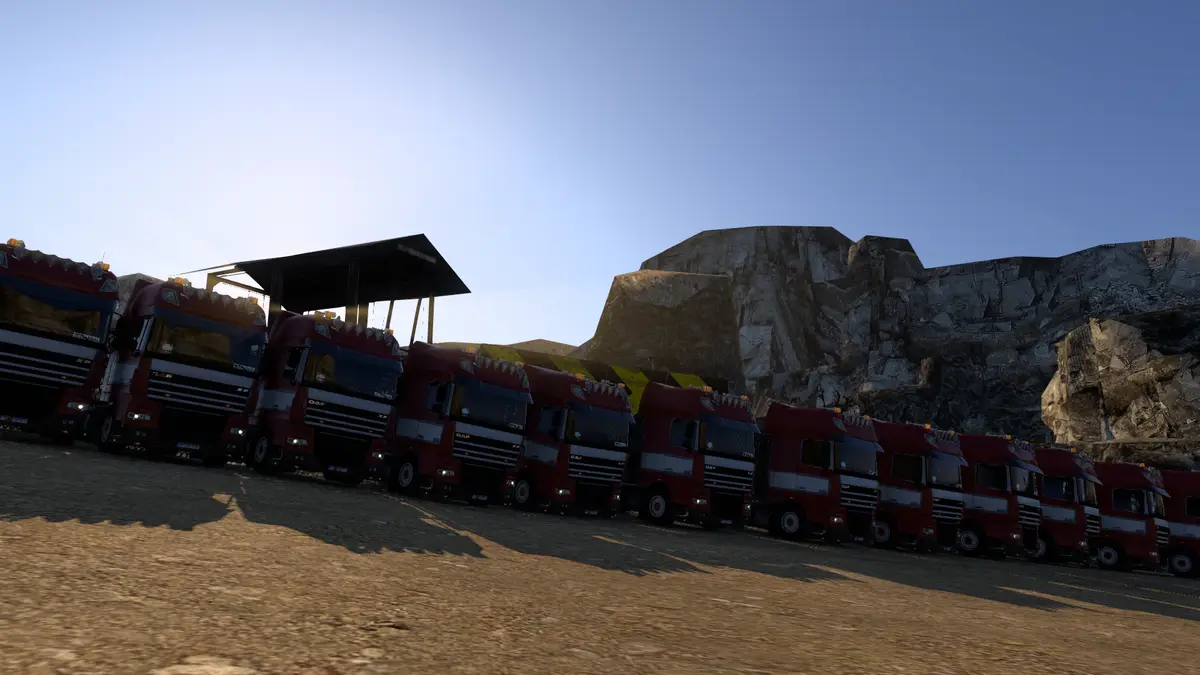 A row of identical red DAF trucks parked in a quarry-like setting under a clear blue sky, with rocky cliffs in the…