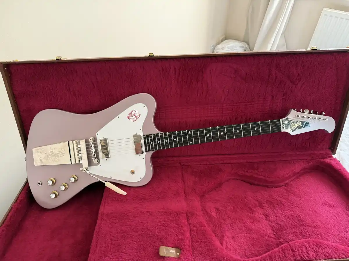 A light purple Gibson electric guitar with a white pickguard and red logo rests inside an open, burgundy-felt-lined hard…