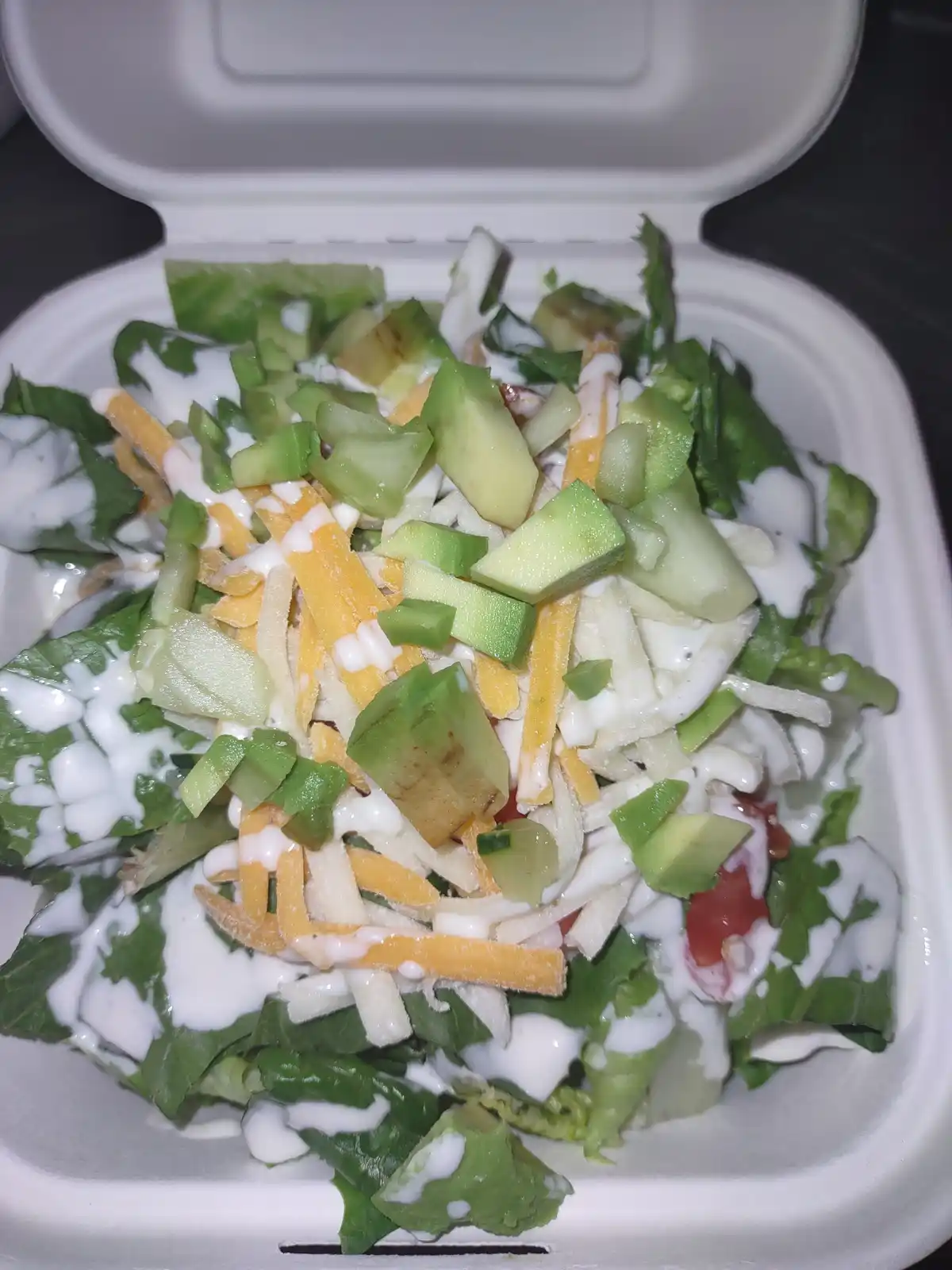 A close-up photo shows a white takeout container filled with a salad topped with avocado chunks, shredded cheese, and a…