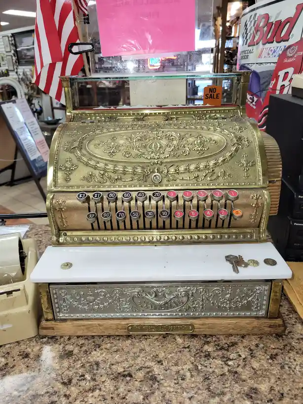 An ornate, vintage brass National cash register sits on a granite countertop in a store, with a small orange 'NO SALE' tag…