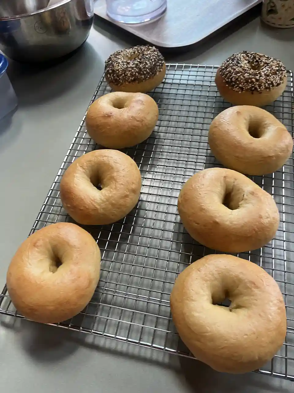 Eight freshly baked bagels, including two seeded ones, sit on a metal cooling rack on a kitchen counter. The overlaid text…