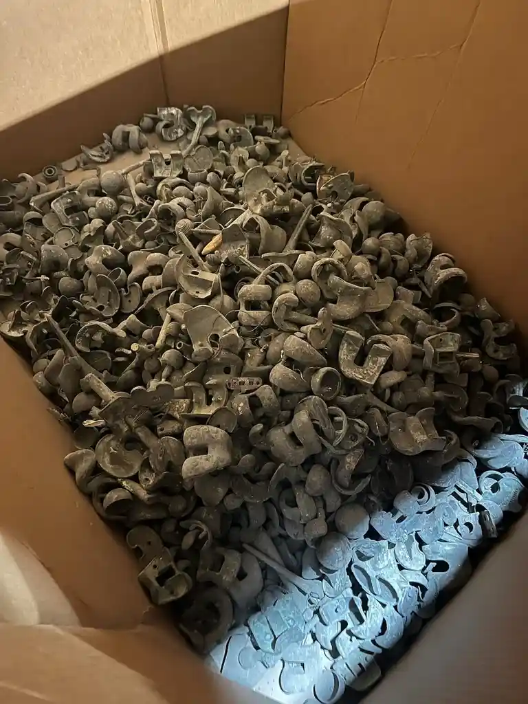 A cardboard box filled with a large quantity of rusty, weathered metal hardware pieces, mostly U-shaped brackets and hooks…