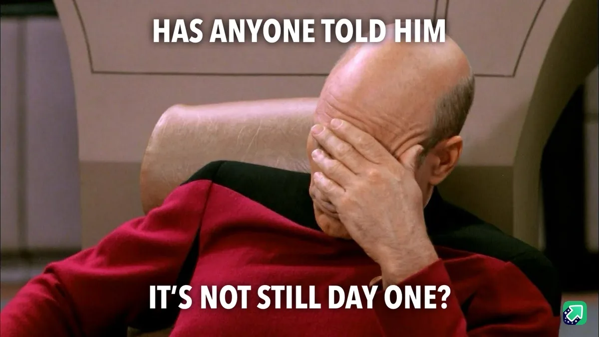 A bald man in a red uniform sits in a chair, covering his face with his hand in a gesture of exasperation or despair. The…