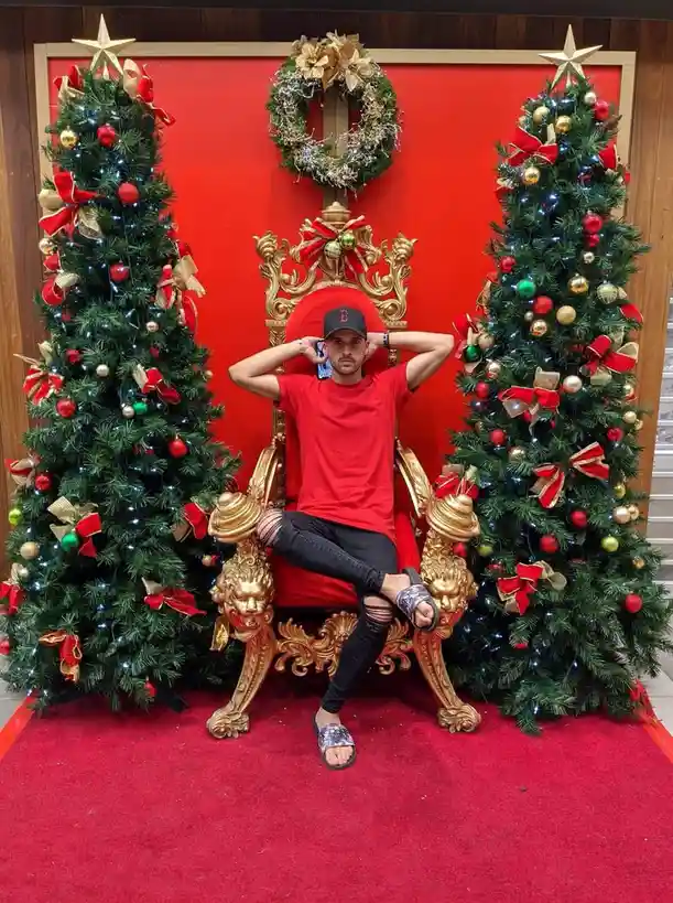A man in a red t-shirt, black ripped jeans, and sandals sits casually on a large, ornate golden throne with red cushions. He…