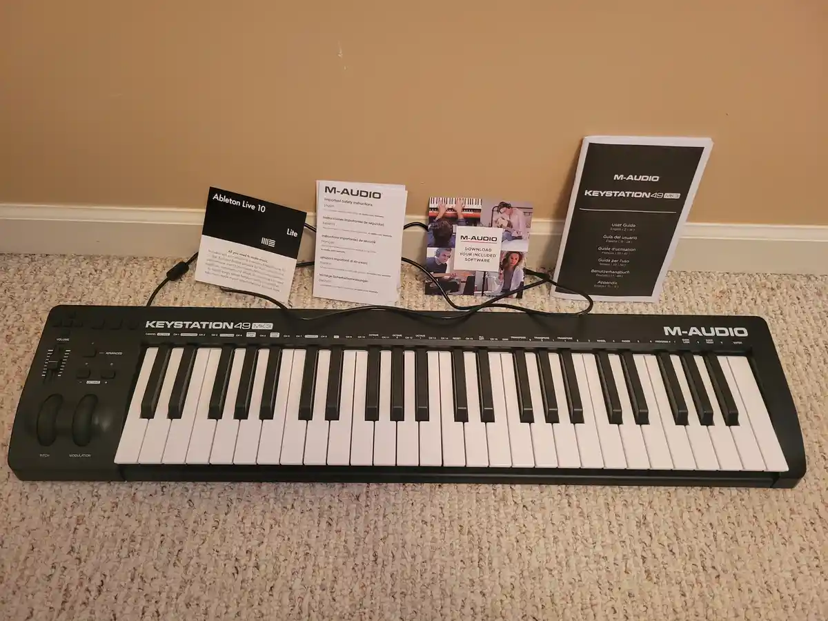A black M-Audio Keystation 49 MK3 MIDI keyboard sits on a beige carpet with its included manuals and cables behind it. The…