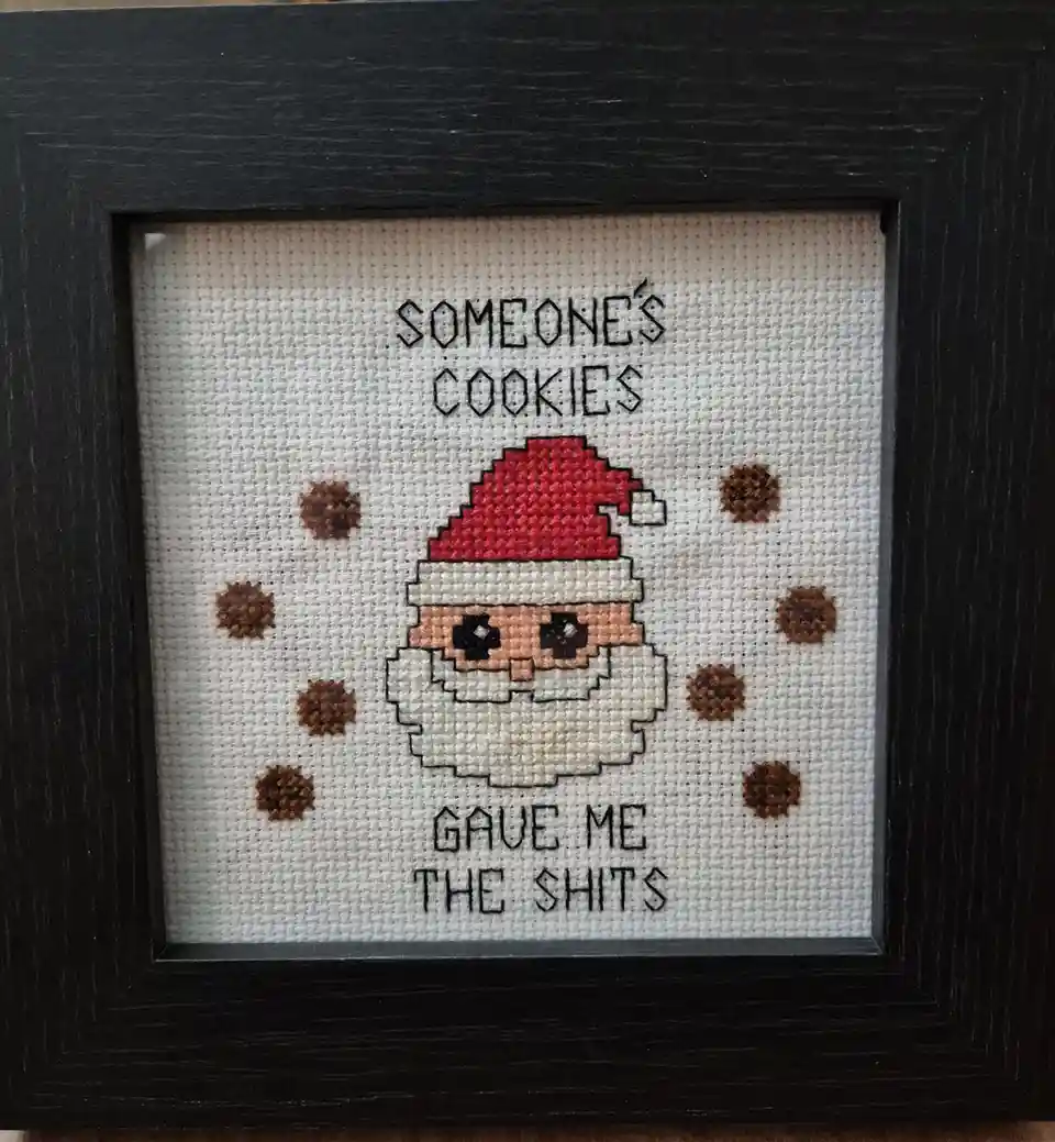 A framed cross-stitch artwork depicts a pixelated Santa Claus with a red hat and white beard, surrounded by brown…