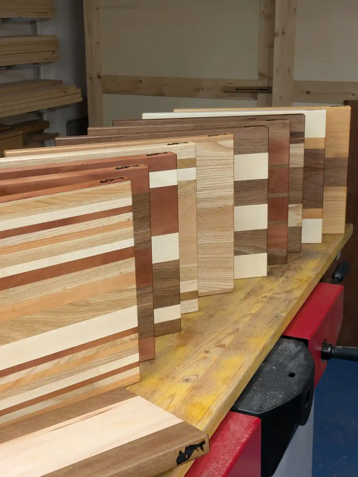 A row of handmade wooden cutting boards with varied striped and solid wood patterns are standing upright on a workshop…