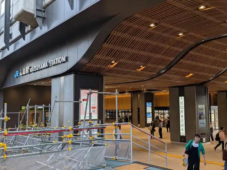 A photo of Matsuyama Station's modern entrance with a wooden slat ceiling and glass walls, partially blocked by metal…