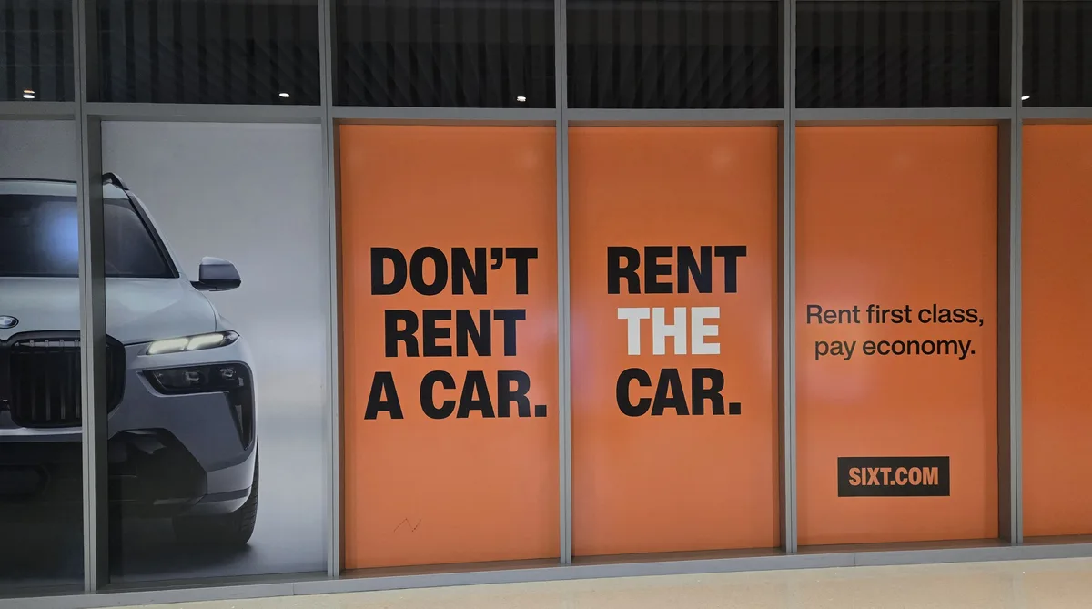 An orange advertisement on a glass storefront features bold black text saying 'DON'T RENT A CAR.' and 'RENT THE CAR.' with…