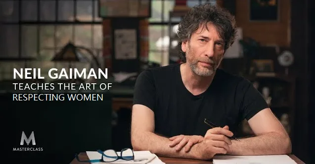A man with dark curly hair and a beard, wearing a black t-shirt, sits at a wooden desk holding a pen. In the background…