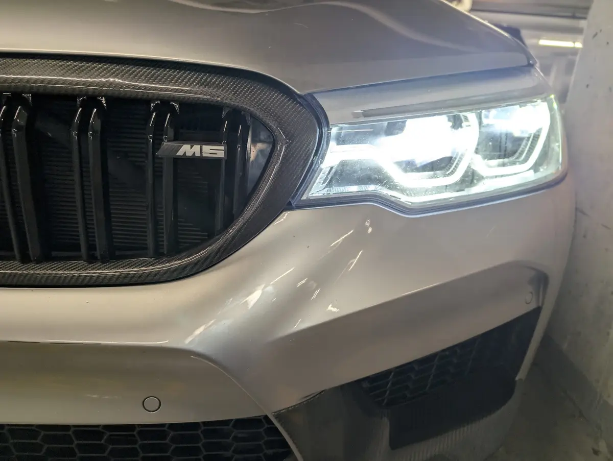 A close-up of the front of a silver BMW M5, showing its illuminated headlight, carbon fiber grille with 'M5' badge, and…