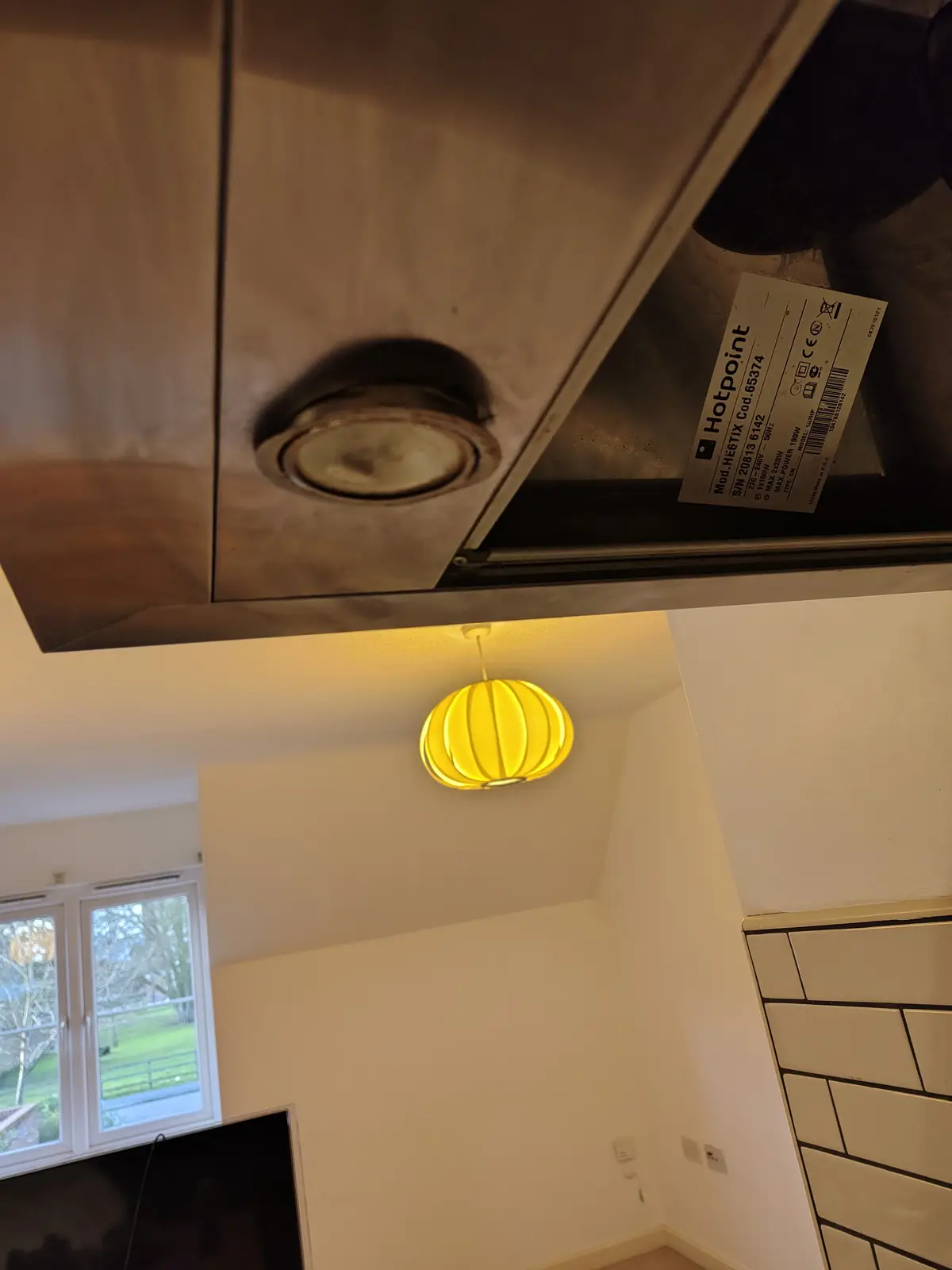 A low-angle photo shows a stainless steel kitchen range hood with a visible Hotpoint brand label, positioned directly above…