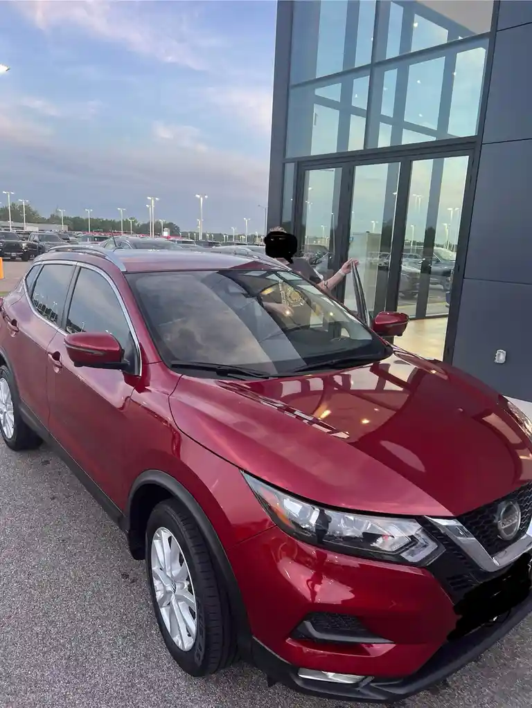 A person with their face obscured stands beside a shiny red Nissan SUV parked outside a modern car dealership with large…
