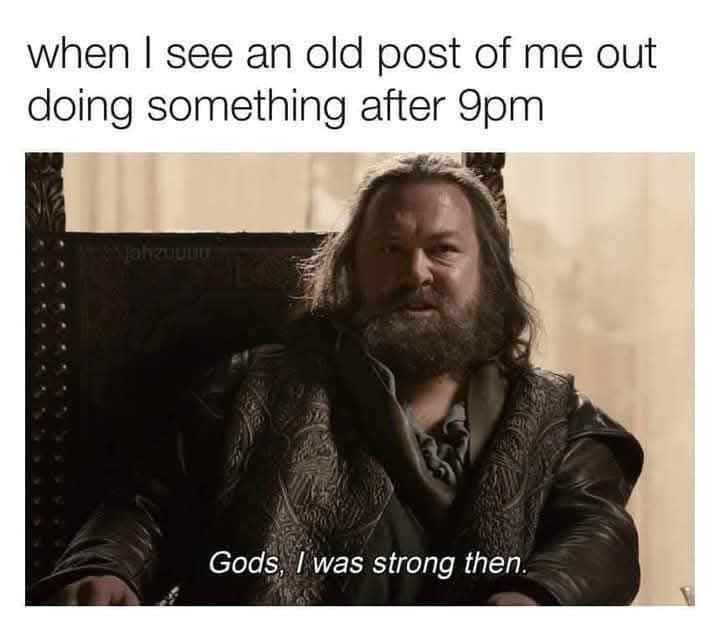 A man with a long beard and dark clothing sits in a throne-like chair, looking back with a nostalgic and slightly amused…