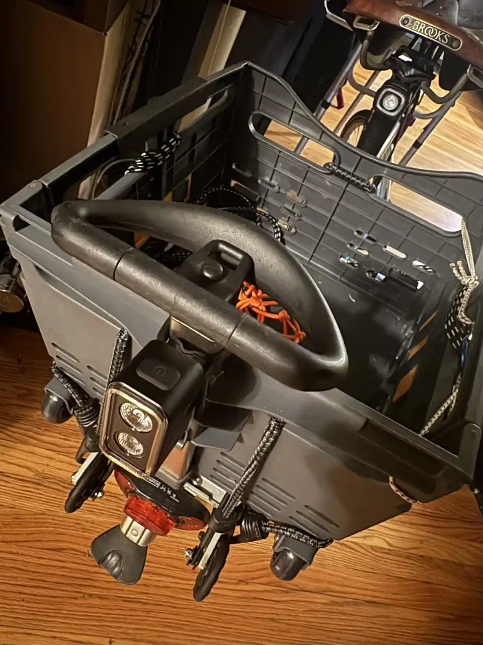A close-up photo shows the front of a bicycle with a large black cargo basket mounted on the front. Inside the basket, a…