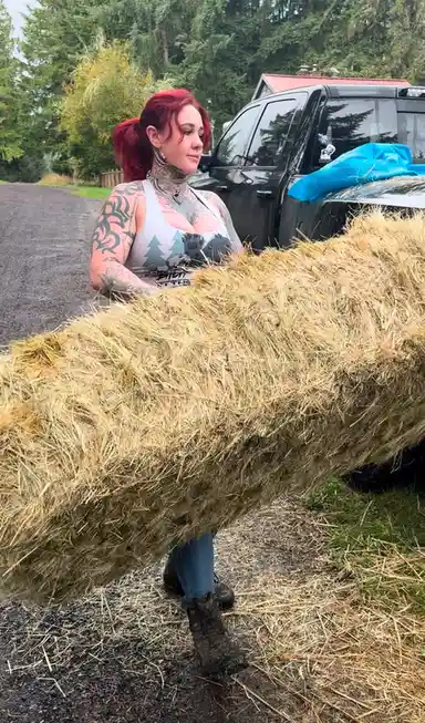 A woman with red hair and extensive tattoos is standing outdoors holding a large bale of hay, which obscures most of her…