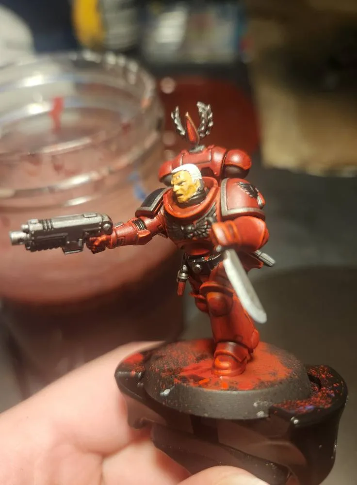 A hand holds a painted red Space Marine miniature, which is posed as if drinking from a glass jar filled with a red liquid…