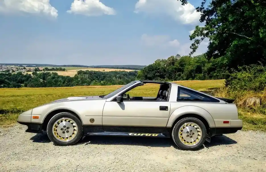 A silver Nissan 240Z with a black stripe and gold-rimmed wheels is parked on a gravel road in a scenic countryside setting…