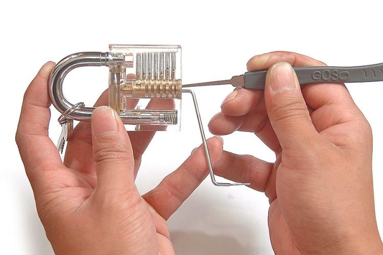 Two hands hold a transparent combination padlock while one hand uses a lock pick tool to manipulate its internal tumblers…